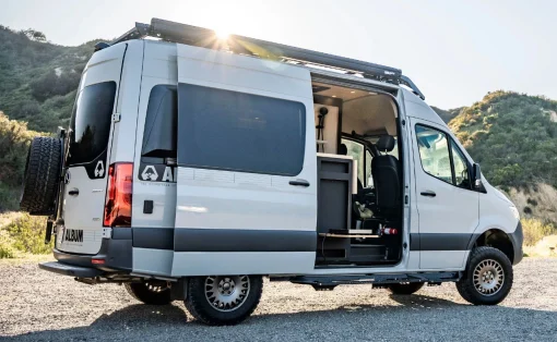 White camper van with its side door open, showing a small living area inside, parked on a gravel lot with hills in the background on a sunny day