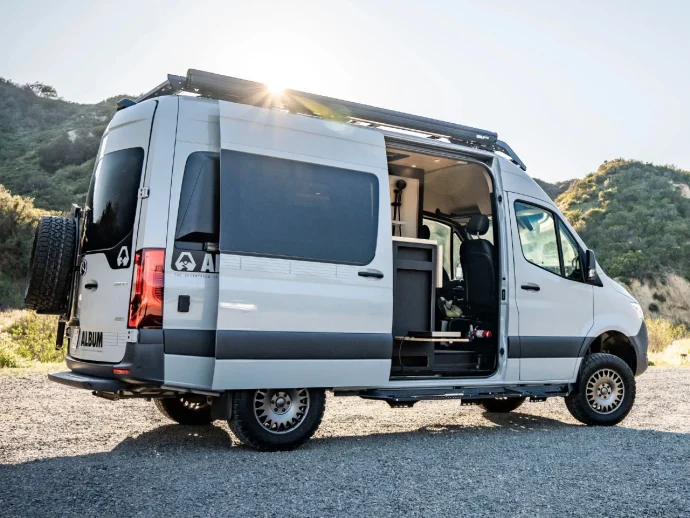 White camper van with its side door open, showing a small living area inside, parked on a gravel lot with hills in the background on a sunny day