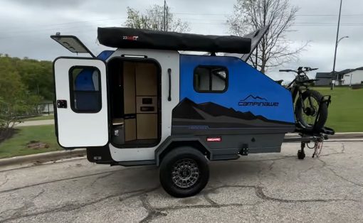 Blue and gray teardrop camper with a mounted bike on the back; side door open revealing a small kitchenette inside; overcast sky.