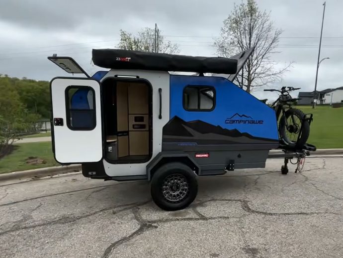 Blue and gray teardrop camper with a mounted bike on the back; side door open revealing a small kitchenette inside; overcast sky.