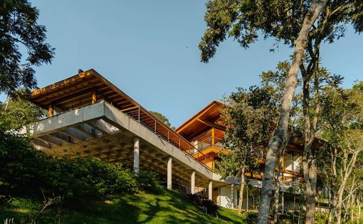 Modern hillside house with exposed concrete pillars and wooden upper levels among trees.