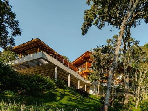 Modern hillside house with exposed concrete pillars and wooden upper levels among trees.