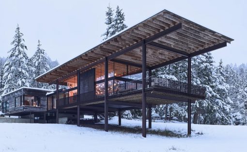 Modern raised house on stilts in a snowy landscape, with glass walls and a large elevated deck, lights glowing from inside.