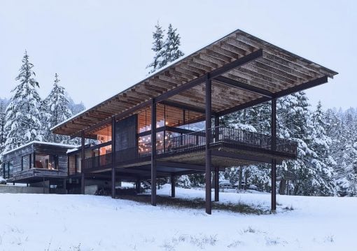 Modern raised house on stilts in a snowy landscape, with glass walls and a large elevated deck, lights glowing from inside.