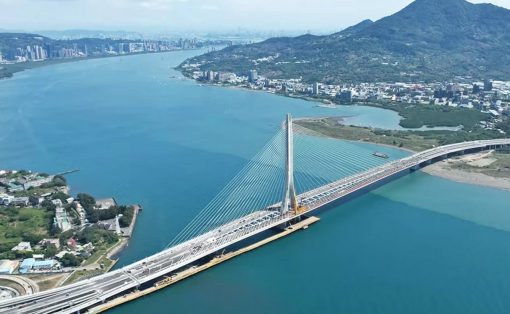 A cable‑stayed bridge crosses a wide blue river, with a city and green hills in the distance.