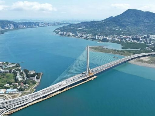 A cable‑stayed bridge crosses a wide blue river, with a city and green hills in the distance.