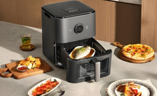 Modern grey air fryer open with a fish fillet inside; surrounding it are dishes like pizza, fried chicken, and a seafood plate on a kitchen counter.