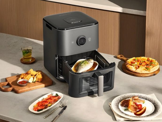 Modern grey air fryer open with a fish fillet inside; surrounding it are dishes like pizza, fried chicken, and a seafood plate on a kitchen counter.