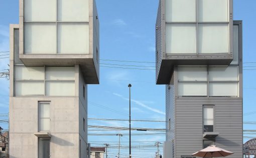 Two cube-shaped, modern buildings with large glass windows sit side by side under a blue sky, with power lines in the background and a patio umbrella in front.
