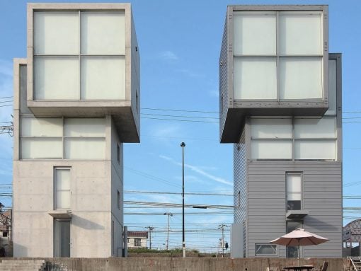 Two cube-shaped, modern buildings with large glass windows sit side by side under a blue sky, with power lines in the background and a patio umbrella in front.
