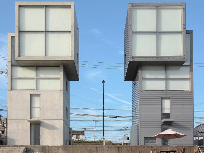 Two cube-shaped, modern buildings with large glass windows sit side by side under a blue sky, with power lines in the background and a patio umbrella in front.
