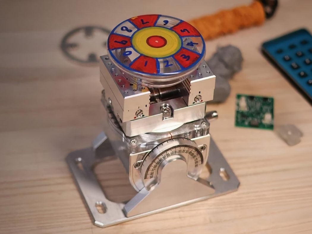 Close-up of a metal mechanical device with a colorful numbered wheel on top, mounted on a silver frame; a circuit board and tools are visible in the background.