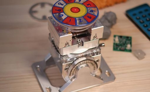 Close-up of a metal mechanical device with a colorful numbered wheel on top, mounted on a silver frame; a circuit board and tools are visible in the background.