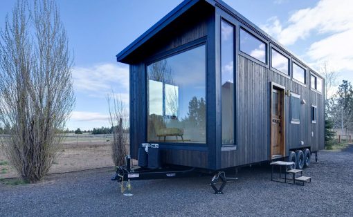 Dark wood tiny house on a trailer with a large glass wall, parked on gravel under a blue sky in a rural setting.