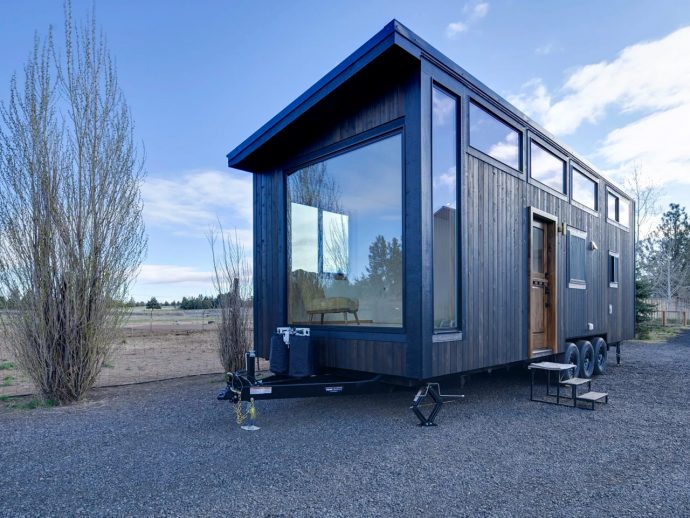 Dark wood tiny house on a trailer with a large glass wall, parked on gravel under a blue sky in a rural setting.
