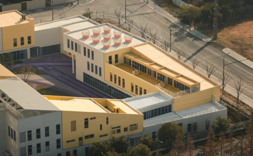 Aerial view of a modern campus building complex in yellow and white, with a purple running track and surrounding roads beyond.