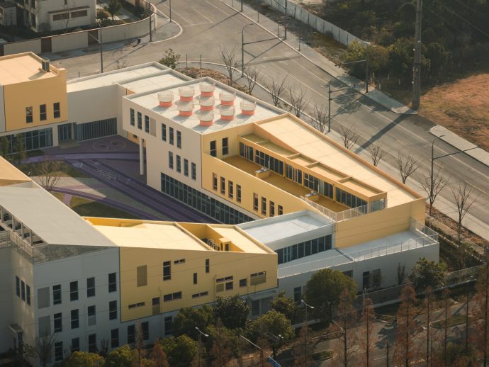 Aerial view of a modern campus building complex in yellow and white, with a purple running track and surrounding roads beyond.