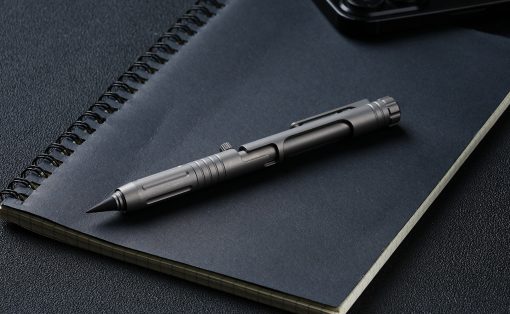 Tactical metal pen resting diagonally on a dark notebook with a spiral binding, on a textured desk surface, with a smartphone nearby in the corner.