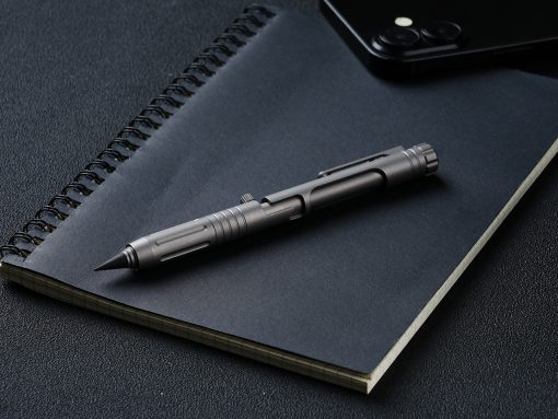 Tactical metal pen resting diagonally on a dark notebook with a spiral binding, on a textured desk surface, with a smartphone nearby in the corner.