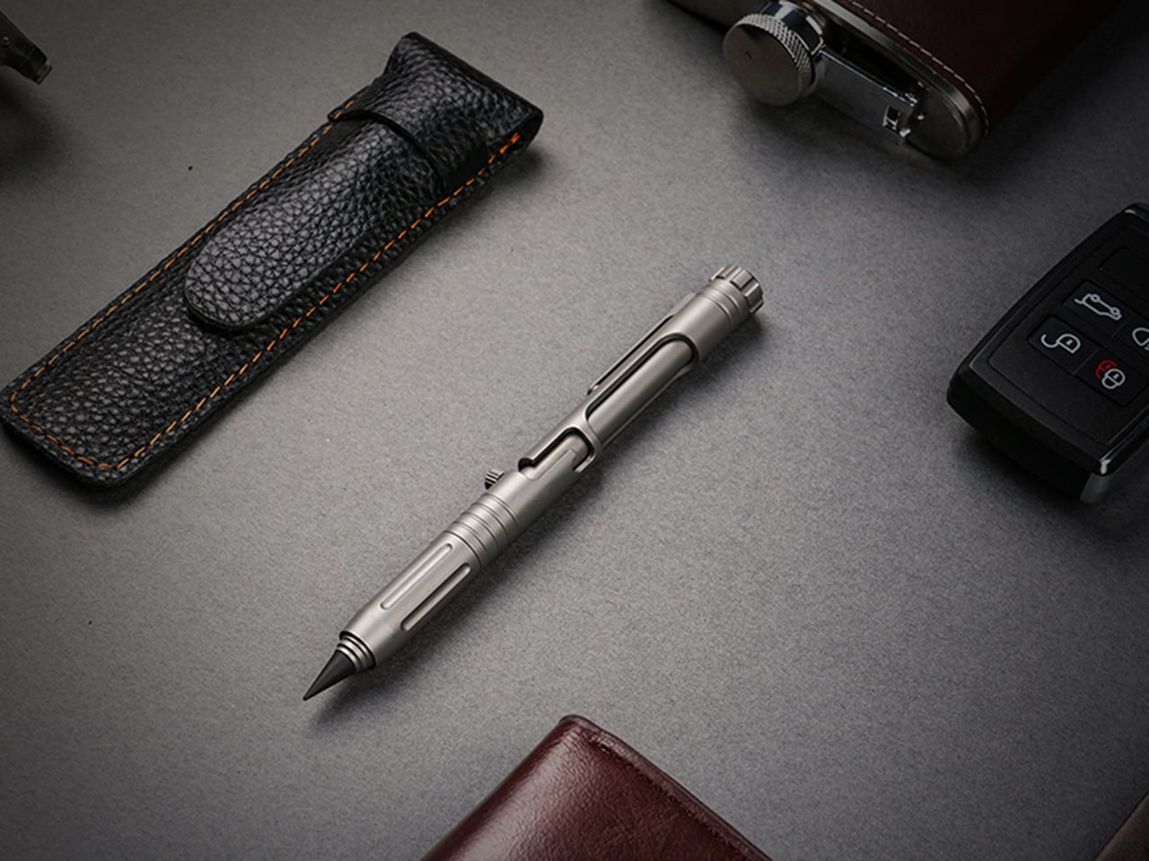 Silver tactical pen on a gray desk with a black leather pen case nearby.