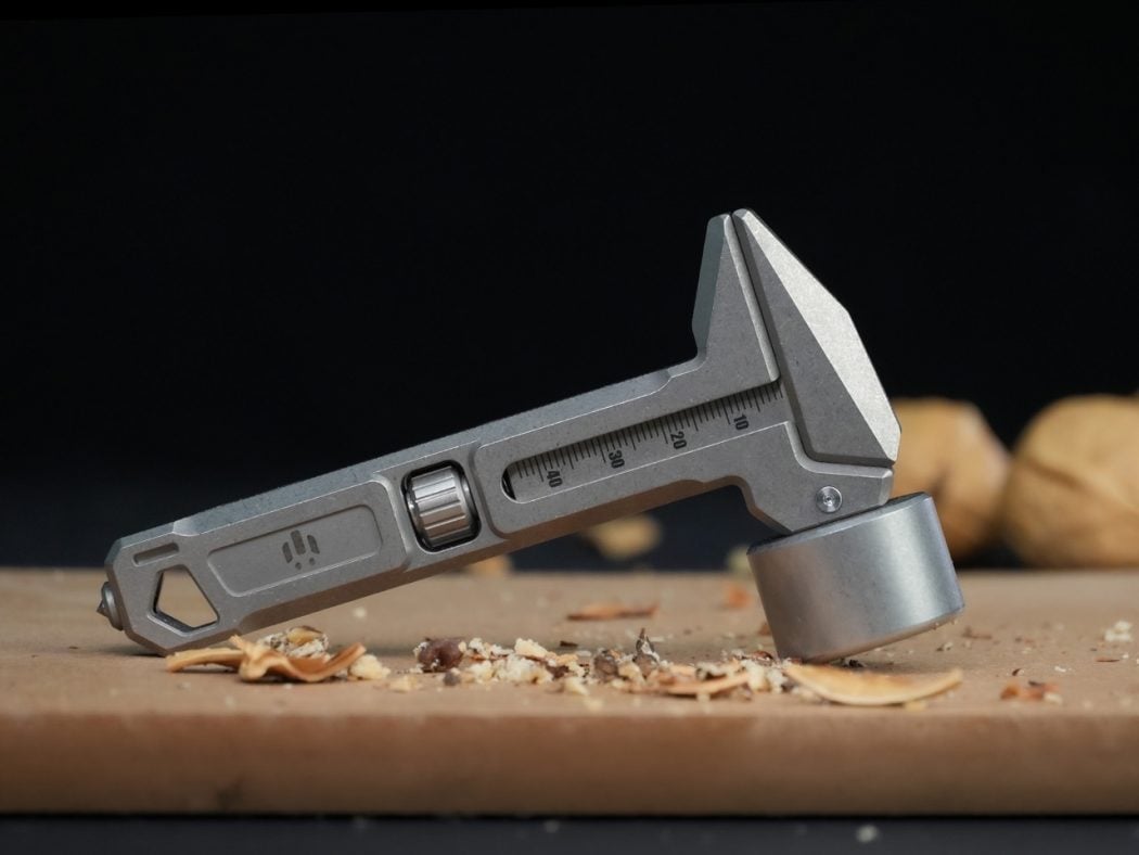 Close-up of a metal caliper with a scale and jaws open, resting on a wooden work surface with wood shavings nearby.
