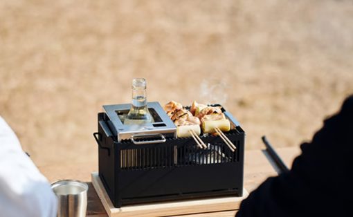 Tabletop charcoal grill with skewered meat and vegetables cooking outdoors, a bottle resting on the grill lid.