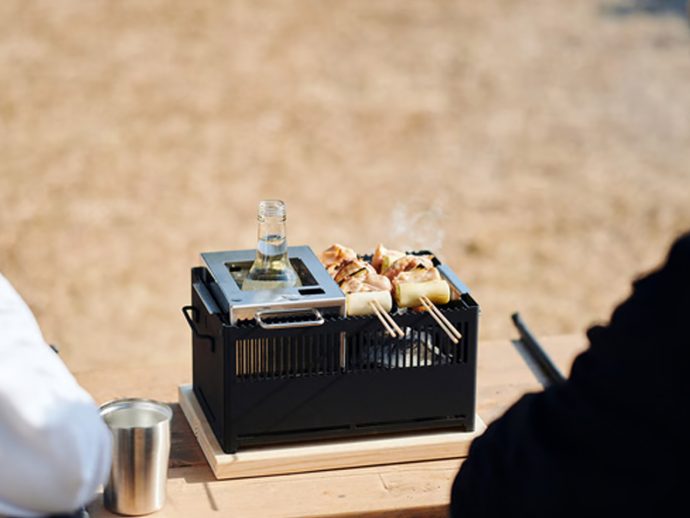 Tabletop charcoal grill with skewered meat and vegetables cooking outdoors, a bottle resting on the grill lid.