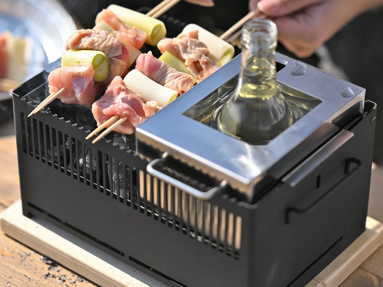 Skewers of meat and green onions grilling on a small portable charcoal grill with a metal insert holding a glass bottle.