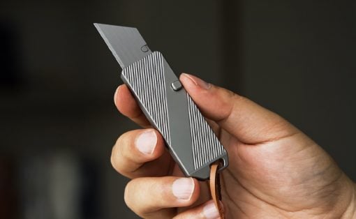Person holds a compact gray multitool with a fold-out blade and striped sides, attached to a brown leather lanyard.