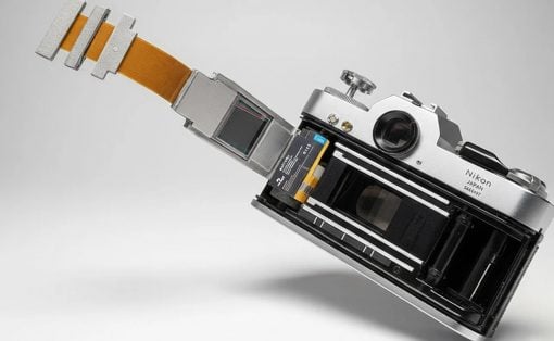 Close-up of a Nikon SLR with the film compartment exposed and an orange flexible cable extending from the back, on a light gray background.