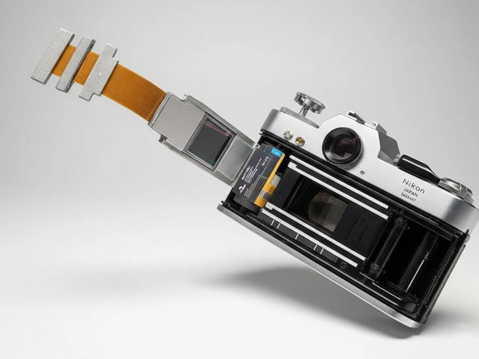 Close-up of a Nikon SLR with the film compartment exposed and an orange flexible cable extending from the back, on a light gray background.