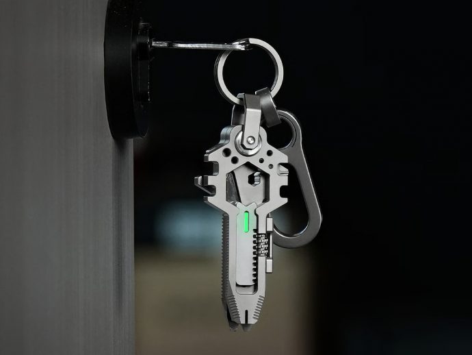 Close-up of a metal multitool keychain with a glowing green indicator hanging from a door knob against a dark background (informative about tool and attachment).
