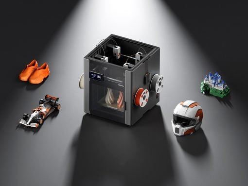 Compact 3D printer under a spotlight, surrounded by orange sneakers, a race car model, a castle, and a helmet to show printing versatility.