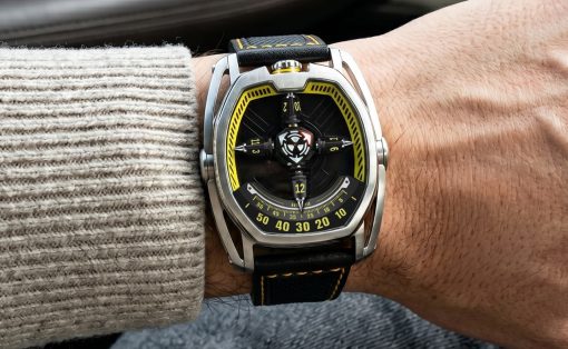 Wristwatch with a black dial and yellow accents on a metal case and black strap, worn on a person's wrist in a knit sweater and jeans.