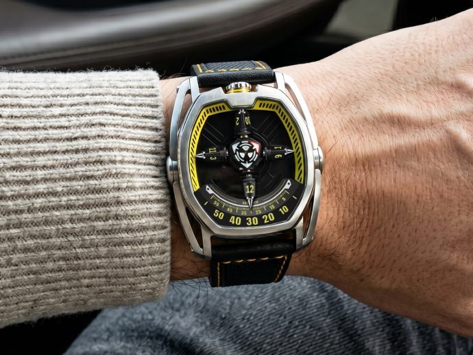 Wristwatch with a black dial and yellow accents on a metal case and black strap, worn on a person's wrist in a knit sweater and jeans.