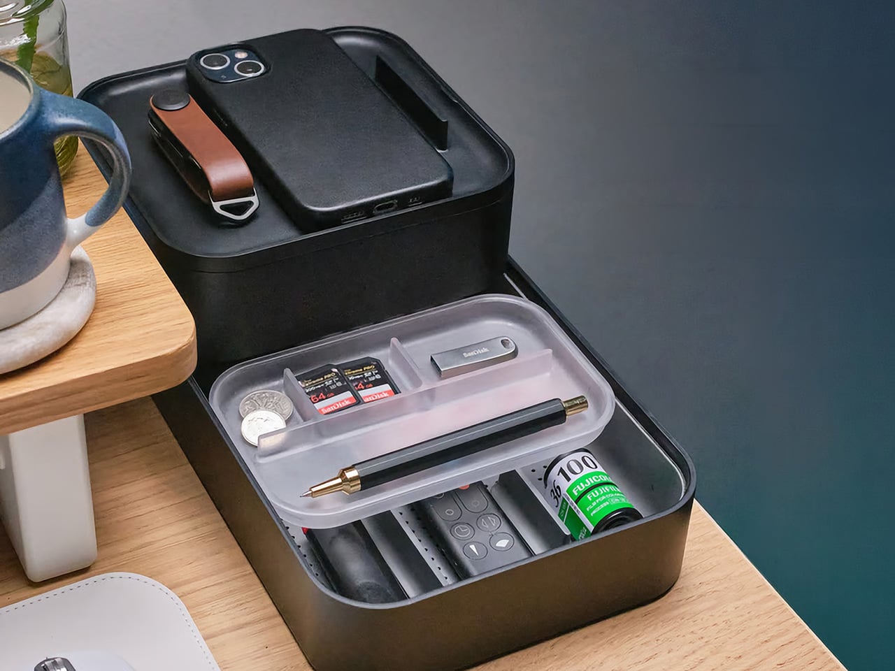 Open black camera/tech case on a wooden desk, revealing small items: memory cards, coins, a USB drive, a fountain pen, and a small bottle with a green label in a clear tray.
