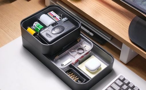 Two‑tier black desk organizer opened to reveal a camera, batteries, tape, and small accessories on a wooden desk.