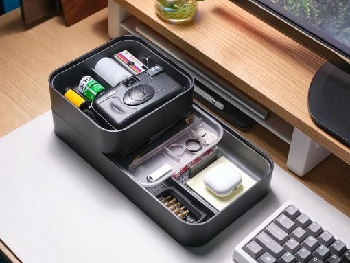 Two‑tier black desk organizer opened to reveal a camera, batteries, tape, and small accessories on a wooden desk.
