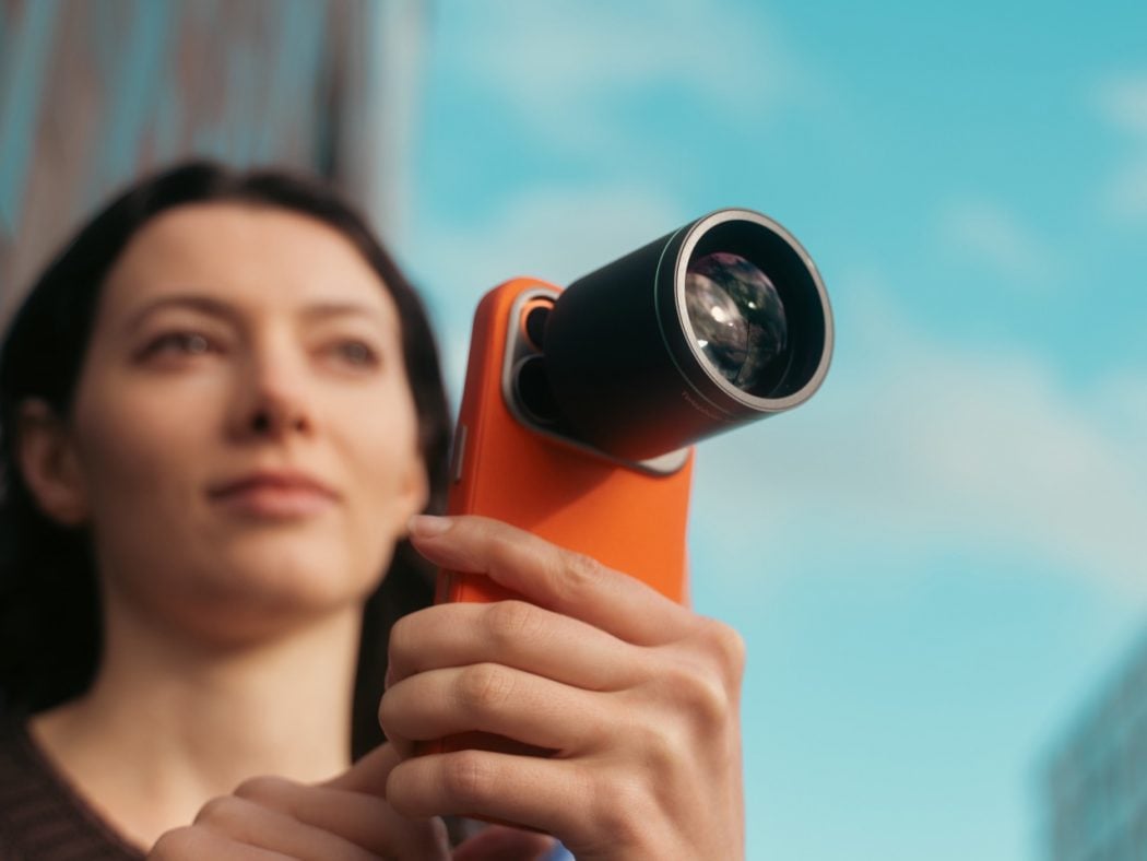 Woman holding an orange handheld device with a large external lens, looking upward against a blue sky.