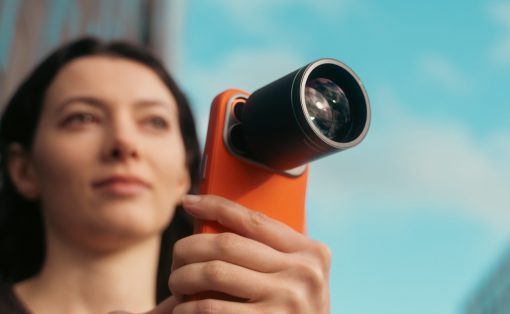 Woman holding an orange handheld device with a large external lens, looking upward against a blue sky.