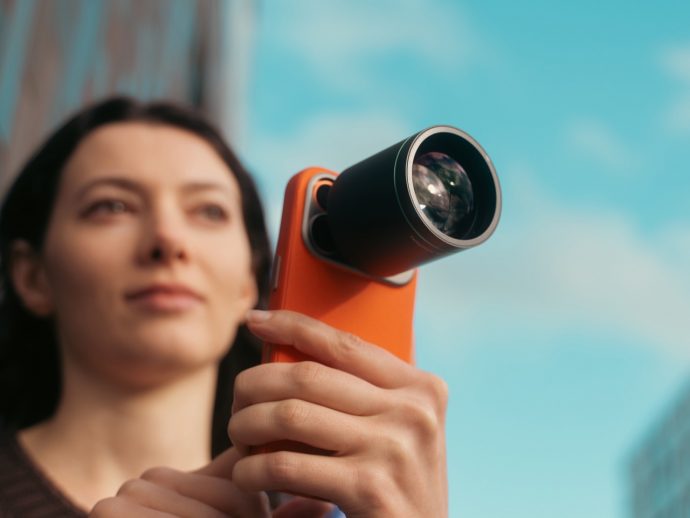 Woman holding an orange handheld device with a large external lens, looking upward against a blue sky.