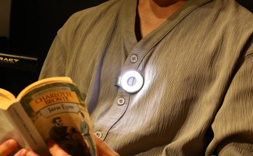 Person reading a paperback titled 'Jane Eyre' by Charlotte Brontë, with a circular clip-on light attached to their chest.