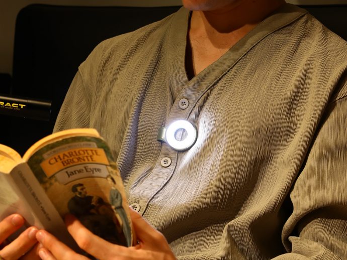 Person reading a paperback titled 'Jane Eyre' by Charlotte Brontë, with a circular clip-on light attached to their chest.