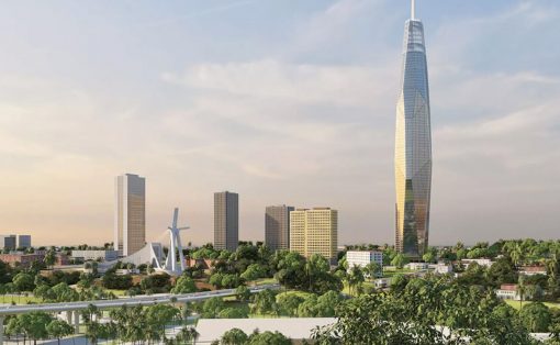 City skyline with a distinctive tall glass skyscraper on the right and low-rise buildings surrounded by trees in the foreground park area.