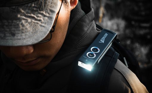 Person in a cap and dark jacket with a small rectangular device clipped to their shoulder strap, emitting a bright light at the bottom.