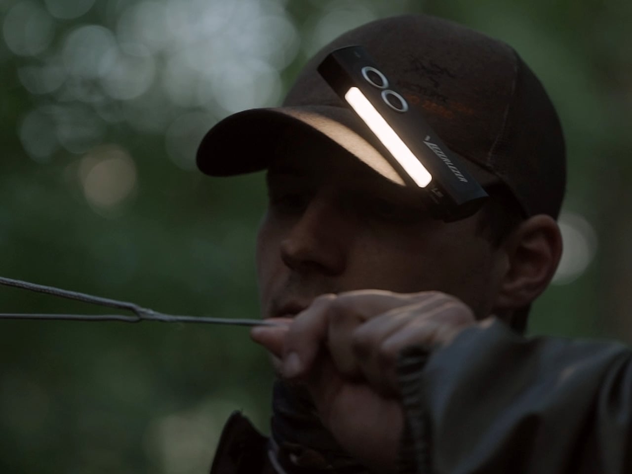 Archer wearing a cap with a mounted headlamp, drawing a bowstring in a dim forest light.