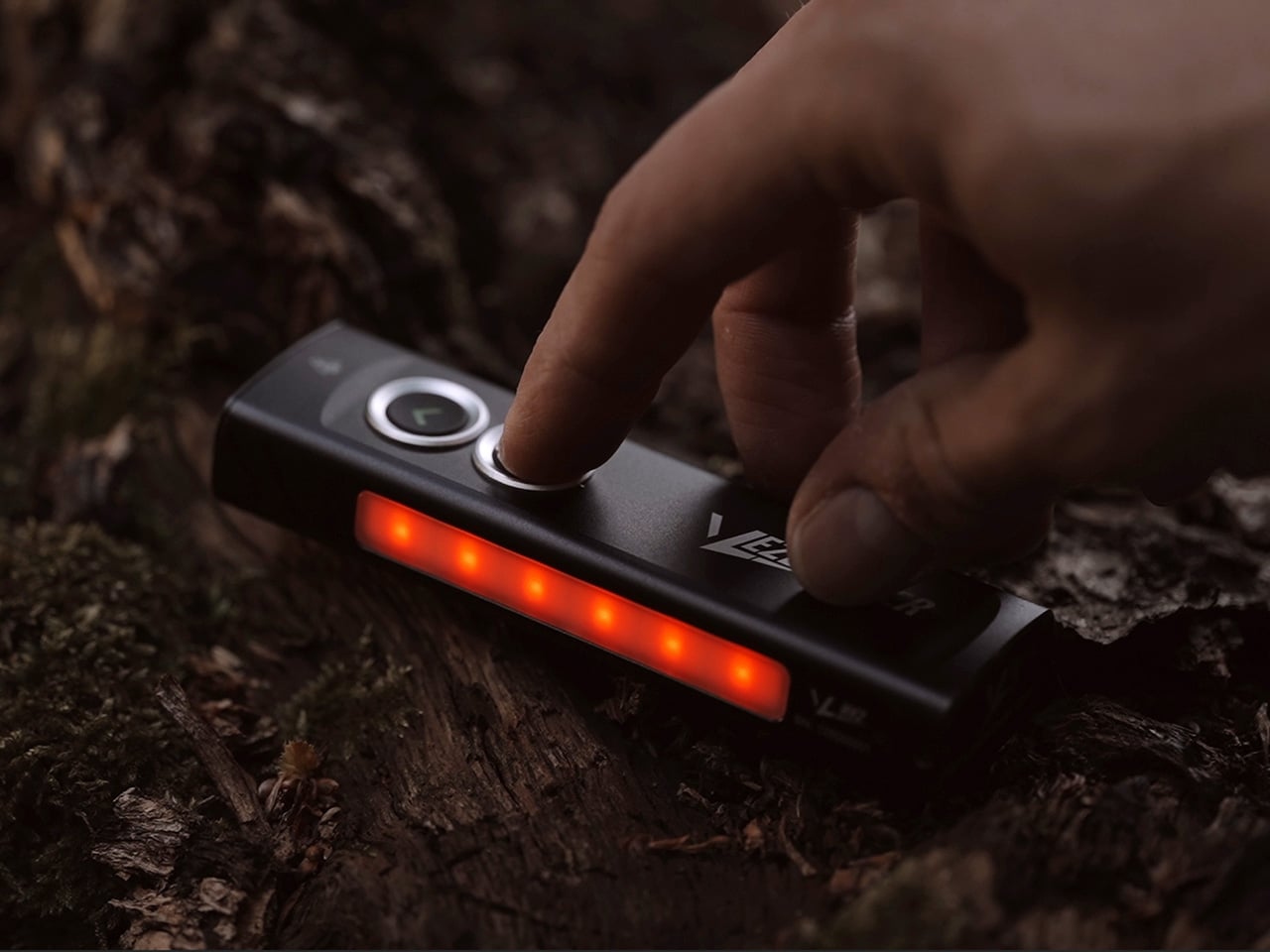 Close-up of a hand pressing a button on a small black device with a red LED bar outdoors on a log.