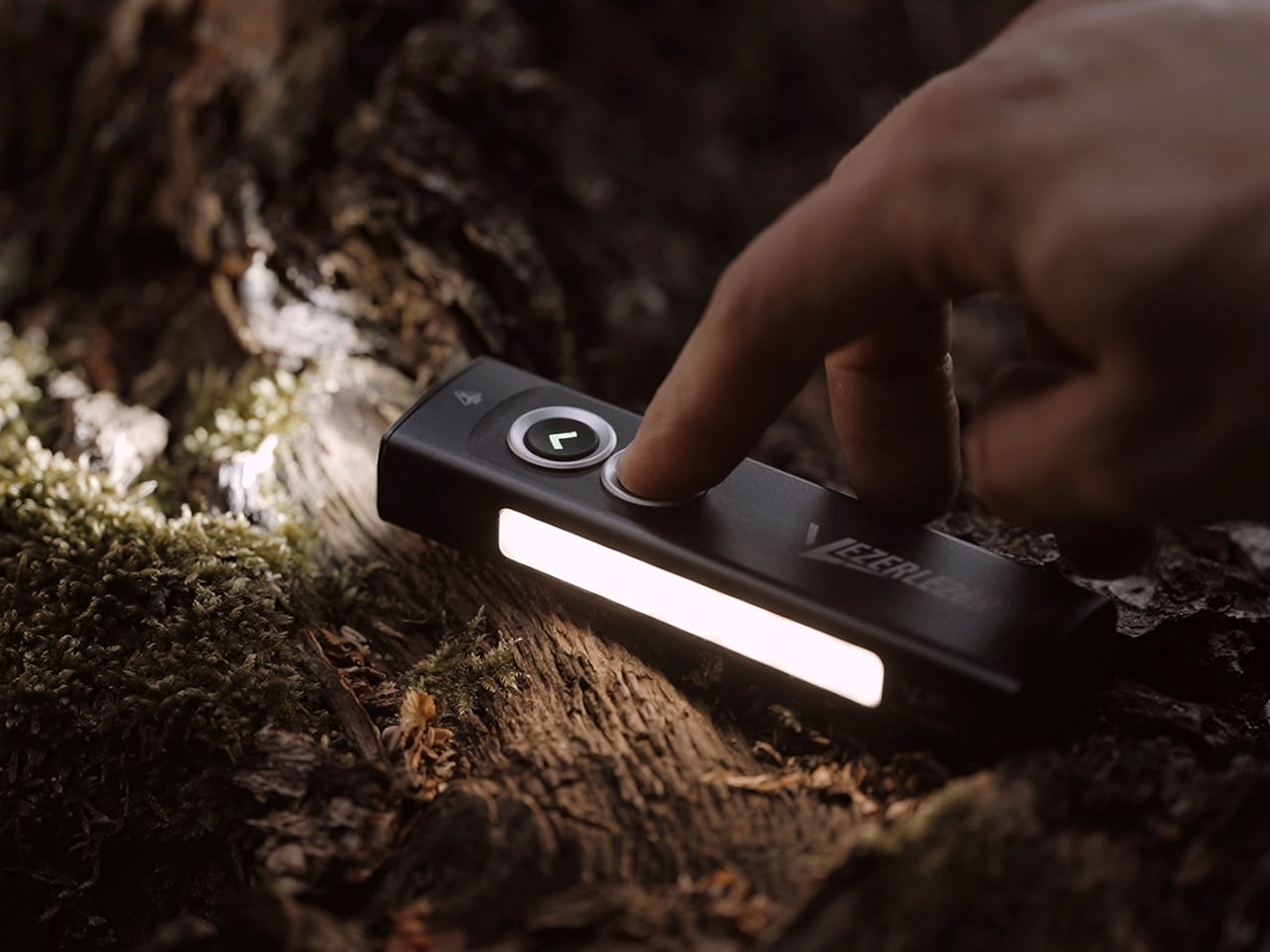 Close-up of a finger pressing a button on a small rectangular outdoor LED light resting on a mossy log in a forest.