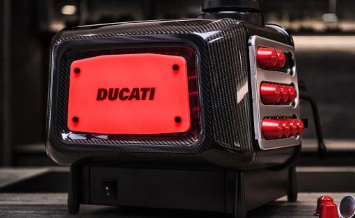 Close-up of a carbon-fiber Ducati portable power unit with a red illuminated panel reading DUCATI on a workshop surface.