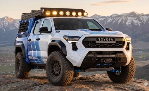 White Toyota pickup with roof rack and eight lights, parked on a rocky overlook with snow-capped mountains in the background.